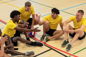Diverse male team in yellow shirts celebrating high fives on gym floor with volleyball, phone