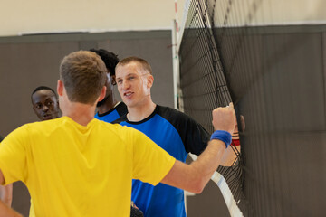 Diverse male volleyball athletes wearing wristband clasping hands through net at gym
