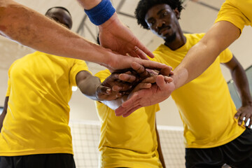 Diverse male athletes stacking hands in huddle at gym court wearing yellow jerseys, black shorts