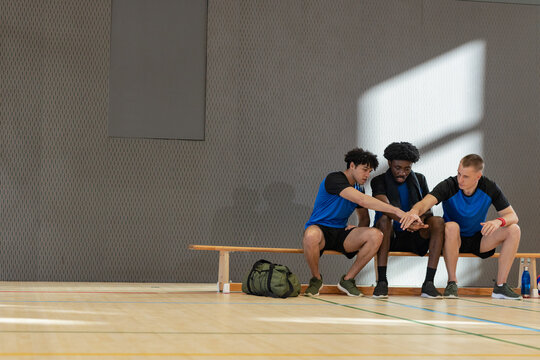 Diverse male players huddling with hands together on bench at gym with duffel bag, copy space