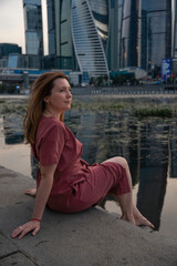 Fototapeta premium Woman in a red dress sits by the Moscow River with the iconic skyscrapers of Moscow City in the background