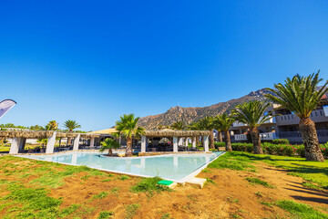 Beautiful view of hotel pool with palm trees, straw roof terrace and mountain view in clear blue sky background. Kos. Greece.