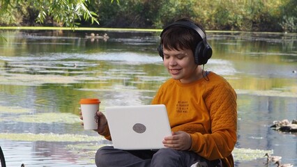 boy using laptop computer, teenager relaxing in park and headphones, listening to music, watching movies, chatting with friends online, studying online on social networks.. Young smiling man outdoors