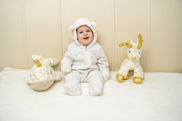 A sweet baby in a cozy white onesie with bear ears sits on a soft bed, looking slightly to the side...