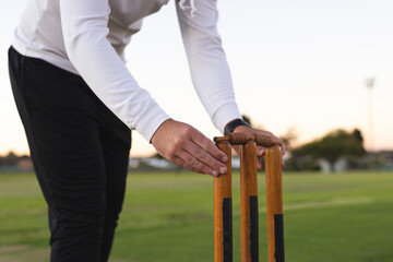 Wooden cricket stumps being adjusted on grassy field by player in white hoodie