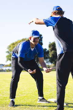 Diverse male teammates in blue caps crouching and signaling on baseball field next to white base
