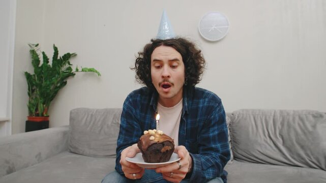 A curly-haired guy sits alone at home, blows out a candle on a cupcake with a sad face. Suddenly, his friends jump onto the couch, surprising him, he lights up with joy