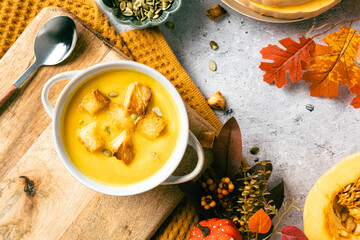 Creamy pumpkin soup with croutons and seeds on a wooden board. Surrounded by autumn leaves, squash,...