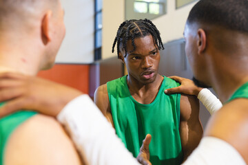 Male basketball teammates in green jerseys forming huddle by padded wall under gym windows