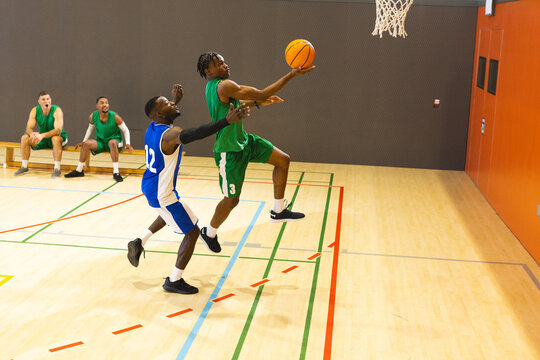 African american male leaping for layup with basketball next to hoop in gym with teammates on bench