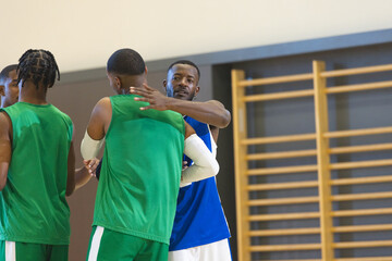 Diverse male basketball players greeting on court wearing green and blue jerseys next to wall bars