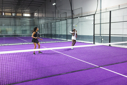 Diverse female sports partners playing padel on purple court with white net, rackets and ball