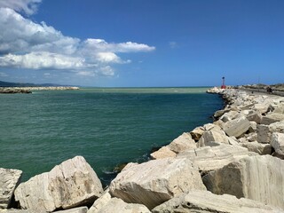 A rocky breakwater on the Adriatic Sea in Italy marks the entrance to a port, with the calm blue-green water meeting a bright blue sky.