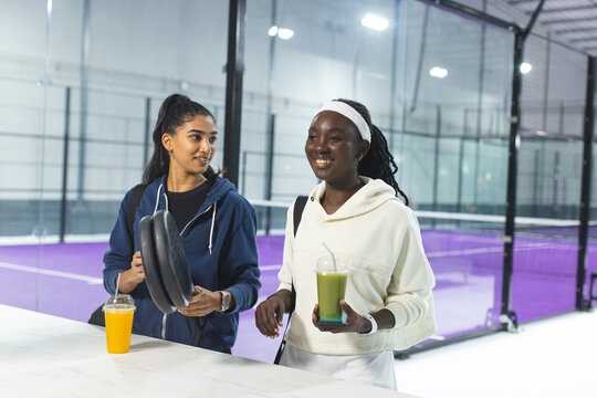 Diverse female friends with padel rackets, smoothies, carrying backpacks at padel facility