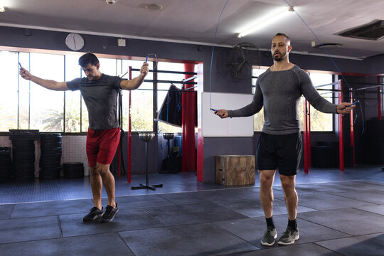 Diverse male workout friends wearing athletic gear skipping ropes in gym featuring weight plates