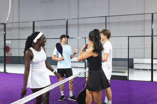Diverse friends standing at padel court net, chatting after match with paddles, fire hose reel