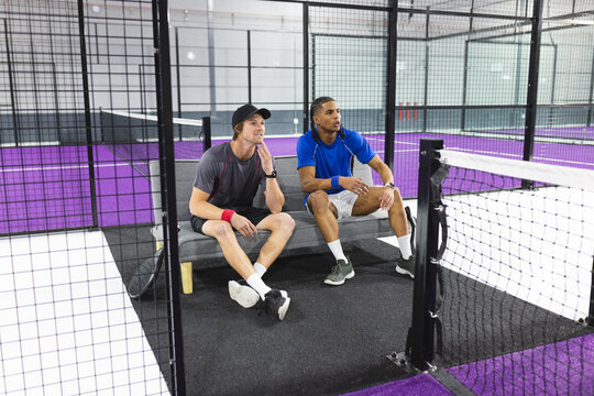 Diverse male friends in sportswear sitting on bench inside padel court beside net, purple turf - Powered by Adobe