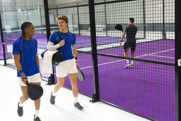 Diverse male athletes in matching uniforms walking padel center with rackets and bag, copy space