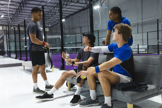 Diverse male friends chatting and resting on padel court bench with padel rackets and drinks