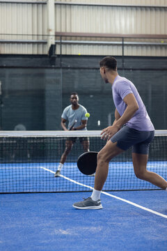 Diverse male opponents swinging rackets on blue pickleball court and hitting ball over net