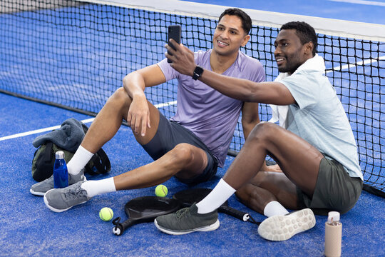 Diverse male friends taking selfie with smartphone on paddle court among paddles, tennis balls - Powered by Adobe