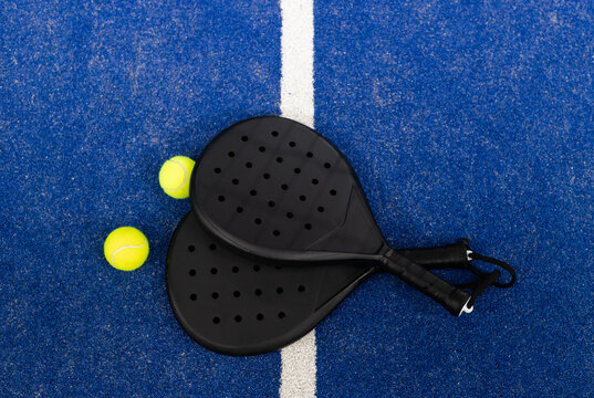 Two black padel rackets overlapping near white court line on blue court with tennis balls