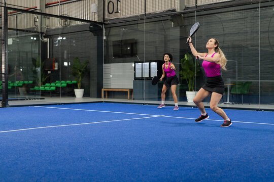 Diverse female teammates playing padel on blue turf court next to net and glass walls, copy space