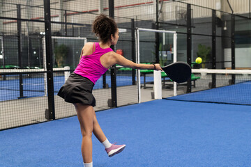 Padel player swinging black paddle hitting yellow ball above net on blue court with fences