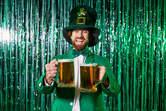 Cheerful man dressed as classic Irish leprechaun holds two frothy beer mugs stand against green glittery party background. St. Patricks Day celebration, Irish pub promotions, festive bar event concept