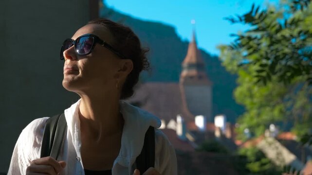 Tourist enjoying sunlight. Female tourist basking in warm sunlight near historic clock tower, wearing white shirt and sunglasses, experiencing relaxed moment within medieval cityscape