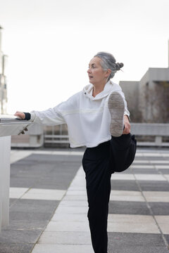 Woman performing standing hamstring stretch using railing on rooftop terrace in athletic wear