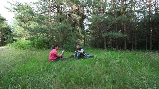 Man and woman resting in a clearing in the forest. Sitting in the grass and drinking water. Family outing, hiking in the forest.