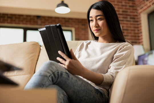 Asian woman using tablet on couch, enjoying flexible remote work in brick wall living room. Female freelancer works from home, researching startup strategies on digital device.