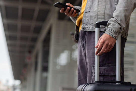 African american man waiting on station platform using smartphone and pulling suitcase