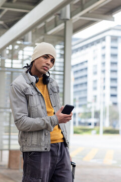 Young man standing at transit shelter holding smartphone and stainless steel travel mug