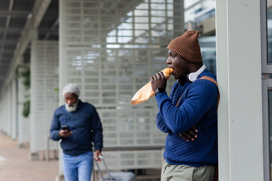 African american male leaning on pillar eating sandwich with headphones under canopy