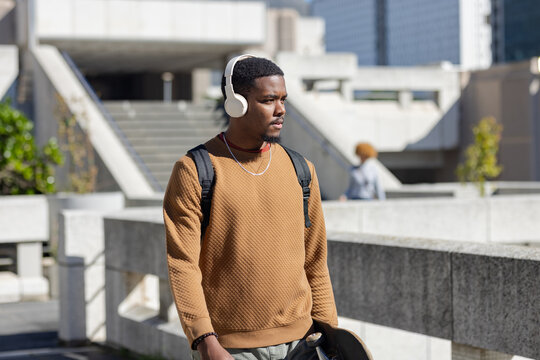 African american man walking in plaza wearing headphones, necklace, carrying skateboard backpack