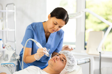 Fototapeta premium Young female cosmetologist performs hardware facial moisturizing procedure to young male patient