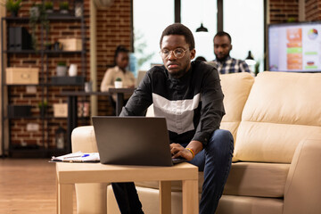 Focused black male manager sitting on couch and using laptop in brick wall office. African american businessman replying to emails and checking daily planner for productive day in modern workspace.