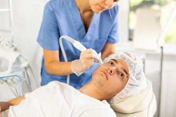 Fototapeta premium Young woman cosmetologist performs facial cleansing procedure with apparatus young male patient