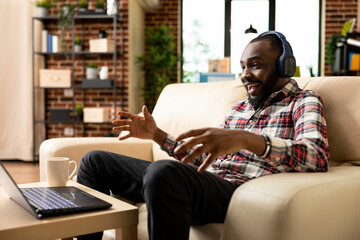 Obraz premium Excited businessman wearing headphones seated at home, enjoying video chat with international work colleagues. Cheerful black man watching movie, streaming content or listening to music on laptop.