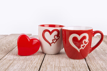 Two heart-themed ceramic mugs and a red heart ornament on rustic wooden surface, symbolizing love, togetherness, and romantic celebration. Ideal for Valentines Day or affectionate lifestyle visuals.