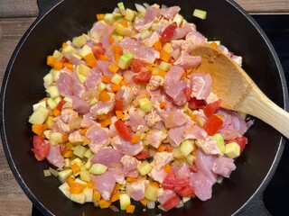 Diced chicken and vegetables are being stir-fried in a cast-iron pan with a wooden spoon. Concept...