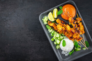 Sate ayamb- grilled chicken satay sate skewers with peanut dipping spicy sauce, steamed rice and fresh cucumber served as top view on tray