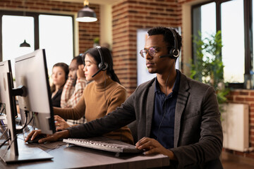 Obraz premium Male company employee wearing wired headphones, focusing on online strategy ideas for digital marketing. African American customer service agent using his desktop pc to review online information.