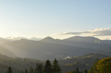Obraz premium Breathtaking view of mountain silhouettes at sunset. The sky is clear, pale blue with few clouds. The sun's rays stream over the peaks, illuminating the landscape below. Carpathian Mountains, Ukraine 