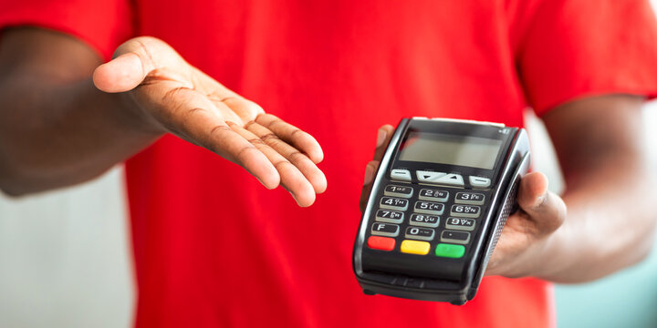 Delivery And Modern Technology. Cropped closeup of black male courier in red uniform holding POS machine in hand, showing payment terminal, blurred background, selective focus - Powered by Adobe