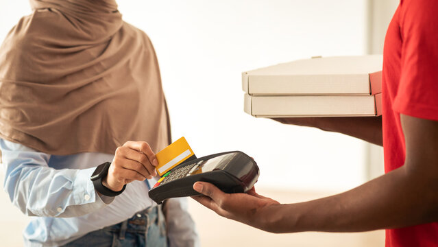 Delivery Concept. Closeup Of Unrecognizable Smiling Islamic Female Customer Paying With Credit Card Standing With Courier In Doorway, Receiving Fast Food Pizza Boxes At Home. Selective Focus On Hand