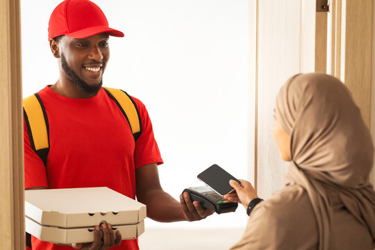 Fast Food Delivery And Cashless Contactless Payment. Smiling black male courier guy wearing red uniform holding Point of Sale terminal and pizza boxes, muslim woman in hijab paying using smartphone