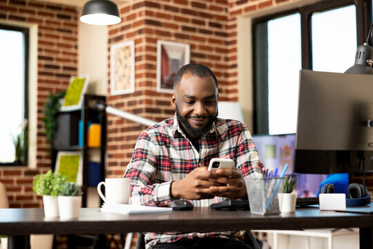 Portrait of happy african american freelancer sitting and using smartphone at modern home office desk. Self employed black man resting from work, networking online using mobile device.